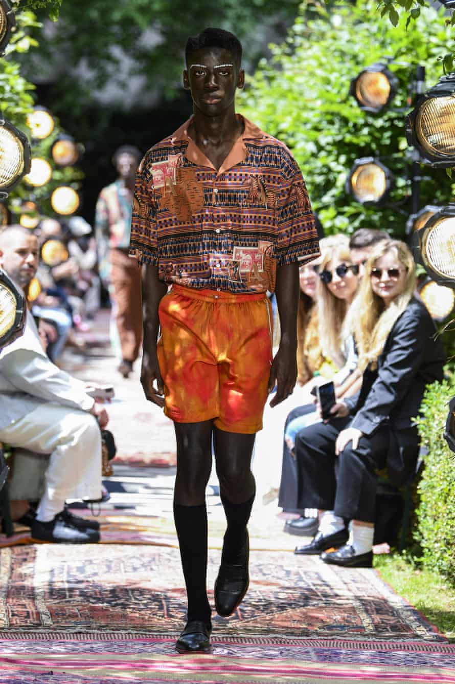 A Black male model is walking down a catwalk, wearing a pair of orange shorts and a short-sleeved, orange patterned shirt, and black shoes and knee-high socks