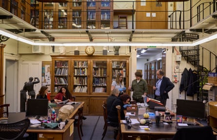 Museum staff work at desks in a back room with winding metal staircases and old wooden bookcases.