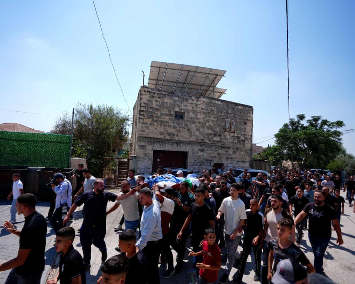 A crowd of men, some of whom are carrying a body on a stretcher that is covered with a sheet, walk along a street