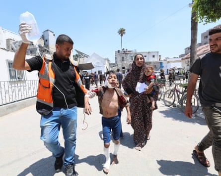 A health worker leads a Palestinian boy whose head and legs are bandaged and holds a saline drip for the boy through a street followed by a crying woman holding a baby