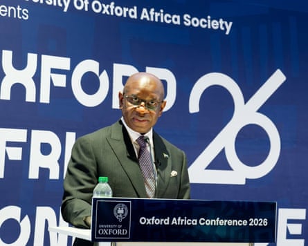 A black man in a suit standing behind a lectern in front of a blue banner emblazoned with white writing