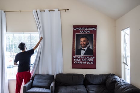 A man opening blinds, a poster of his high school senior picture celebrating his graduation hangs on the wall by the window