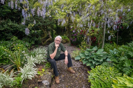 Tim Penrose sitting on a low stone border on a path through a garden with hostas and grasses and wisteria in the background