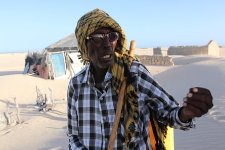 An old man in sunglasses and a turban gestures. In the background are ruined houses half-buried by sand|445x296.66666666666663