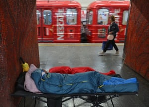 A person wrapped up in a blue sleeping bag in a metro station as a woman walks past on the platform and red train carriages speed by