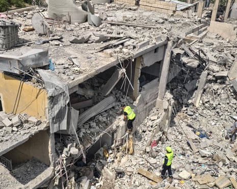 Two men in hi-vis climb on rubble of a destroyed building.