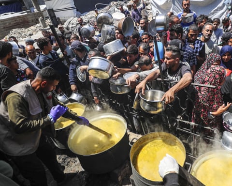 Crowds carrying saucepans and other containers struggle to get them filled from big vats of hot food stirred by people with long pieces of wood