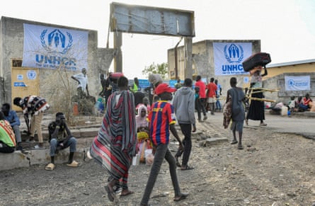 men, women and children carrying baggage walk towards and through a gateway; to each side of it are large white posters with UNHCR logos and lettering.