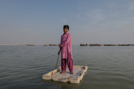 A boy stands on a large piece of polystyrene that may have been the lid of a cool box, holding a stick he is using to punt with.