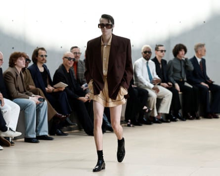 A model in short shorts, a silk shirt, blazer and sunglasses walks down a catwalk in front of spectators