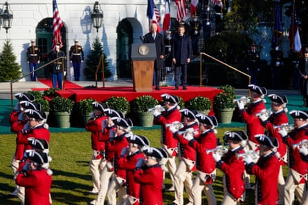 Soldiers dressed in red coats and 18th century military uniforms parade in front of a stage where Joe Biden and Emmanuel Macron are standing.