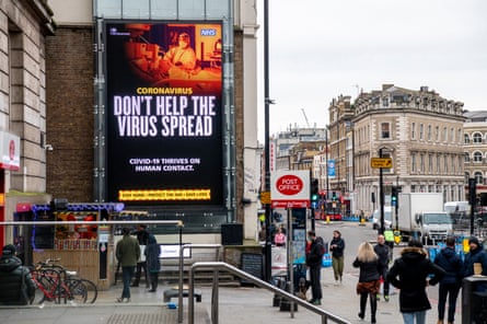 A large billboard sign reads: Act like you’ve got it: don’t help the virus spread. People are walking past in the street and there are some vehicles on the road.