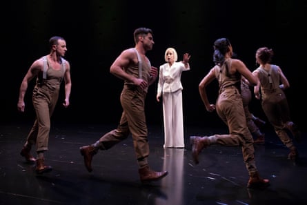 A woman in a white pantsuit with men and women in militaristic overalls running around her in a circle