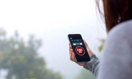 The back of a person, face not visible, holding a smartphone in what looks like a park. The phone is open on a screen saying “place bet”.