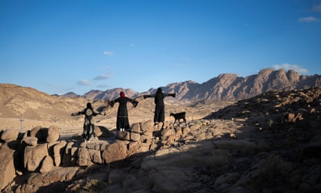 Three women and a goat stand with their arms outstretched on a mountain ridge in the desert
