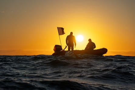 A sunset in the background and a dinghy on the ocean with two people on board.