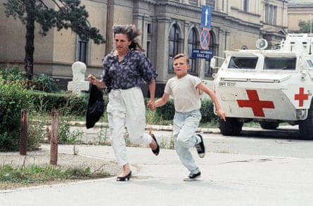 she has long blond hair and wears white jeans and black high-heeled shoes, while her son wears pale blue jeans and a white T-shirt; they are running along a pavement with a stone building in the background and white armoured vehicles with large red crosses on them behind.