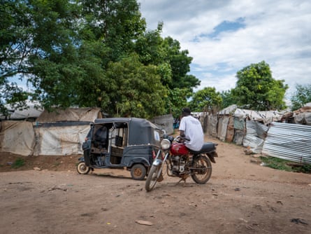 A man sitting on a motorbike next to a tuk tuk on a dirt road lined with tents