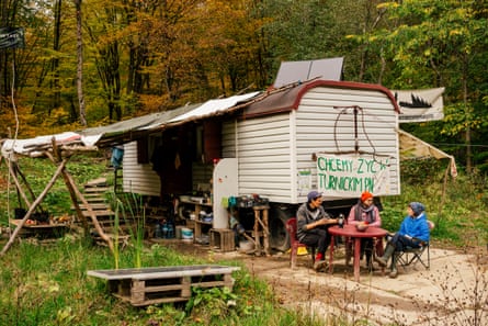 A wooden caravan with an awning in a forest clearing and three people sitting outside