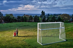 Two children stand near a soccer goal on top of a hill in a beam of light, looking out at a cloudy blue sky.