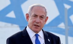 Middle-aged white man, big head, dark suit, blue tie, speaking in front of Israeli flag.