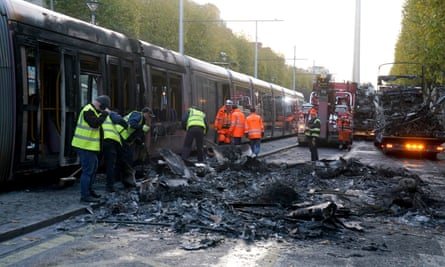 Workers in hi-vis jackets clear debris from the road alongside a fire-damaged tram