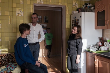 Three members of the family are standing together in the kitchen and the fourth is in an adjoining room seen through an open door