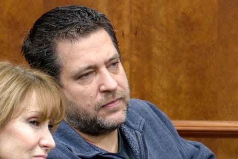 A middle-aged white man with brown hair and a brown beard, wearing a blue zip-up flannel, sits in court.