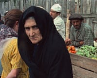 An elderly woman in a black headscarf looks toward the camera at an outdoor market with vendors and vegetables, in a scene from Imperium