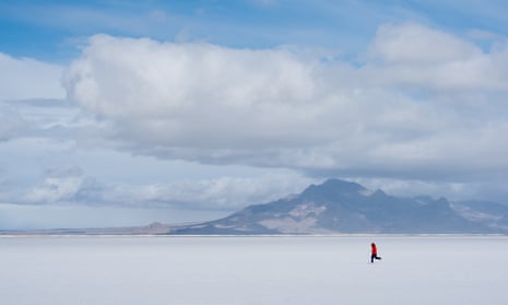 A small figure dressed in a red jacket running across a flat landscape with mountains in the background