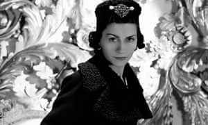 Vogue 1937: Coco Chanel wearing one of her designs: a three-quarter coat with cinched waist and astrakhan lapels and cuffs, and matching astrakhan hat with large brooch on front.