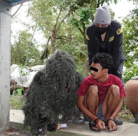 A Thai police officer in a costume that covers every part of his body in what looks like dry grass, next to a figure in handcuffs and another officer