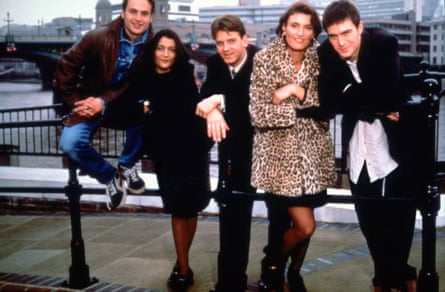 The cast stand against railings on the bank of the Thames with buildings in the background.