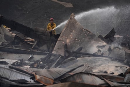 A firefighter in a yellow coat and red helmet sprays water over a pile of charred rubble.