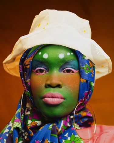 A person with green face paint, a white bucket hat and patterned headscarf gazes forward