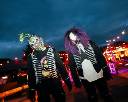 A man in a green face masks stands next to a man in a purple wig as they both tilt their heads to one side and stare scarily.