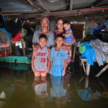 a family of five face the camera standing knee-deep in floodwaters in their own home