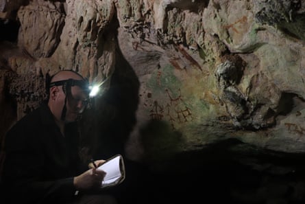 Man with head torch on in a cave writing something down next to a cave painting