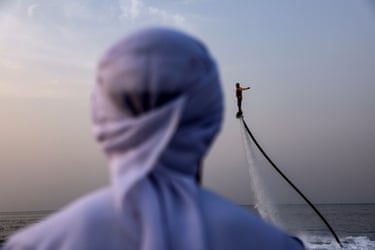 A flyboarder hovers high above the sea in Oman, with a hooded figure blurred in the foreground