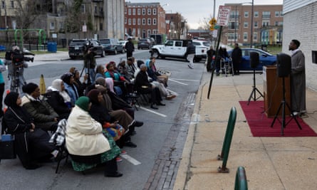 Two long rows of chairs on parking spaces on a city street hold mostly Black people, with a Black man standing at a lectern on a piece of red carpet on the sidewalk in front of them.