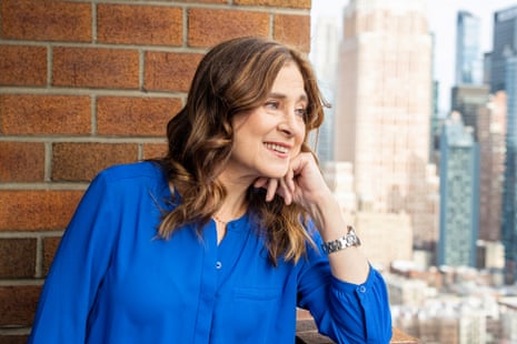 brown haired woman wearing a blue tunic looking out toward the city with a brick wall in the background