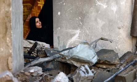 A woman in a headscarf looks out from the window of a house in Rafah with a carpet behind her and concrete rubble and pipes outside