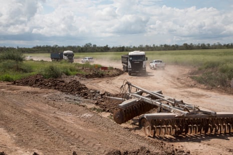 Lorries and pickup trucks drive to and from a half-built dirt road with machinery for clearing land in the foreground