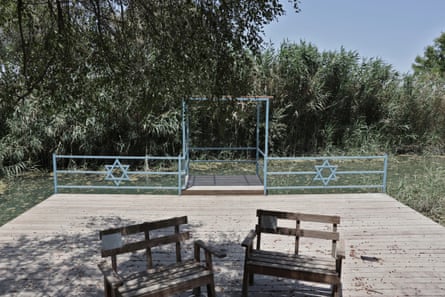 Two benches are placed on decking in the foreground. Behind are metal fences with two Star of Davids part of the metalwork. A pool of water is behind the fence, surrounded by bushes and small trees