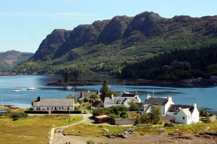 A small cluster of white houses with grey slate roofs sit at the edge of a loch with mountains in the distance