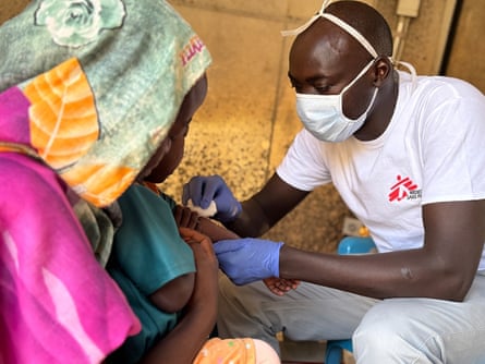 A woman with her back to us holds a child on her lap while a man wearing a face mask touches the child’s arm with cotton wool