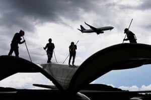 Workers clean a roof at an airport as a plane flies overhead