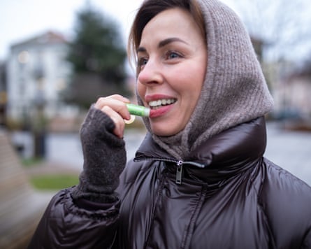 Woman Applying Lip Balm OutdoorsA woman applies lip balm while outdoors on a chilly day. She’s wearing a dark brown hooded jacket and gray gloves.