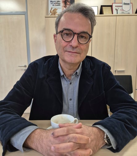 Lucian Mîndruță sits at a pale wooden desk with his hands around an empty white mug. He is in his late 50s with short swept-back grey hair and black-rimmed glasses. He wears a dark jacket and grey open-necked shirt. There are cupboards in the background and he appears to be in an office.