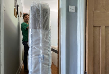 A man stands with the mattress in its wrapping on a landing outside a bedroom door.